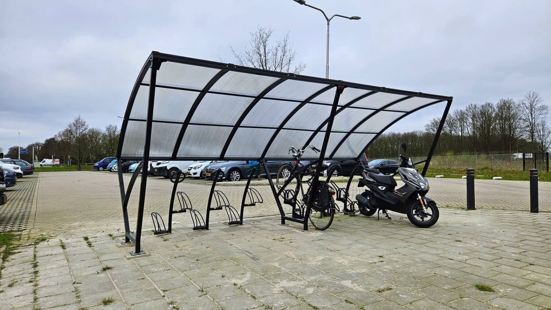 Fietsoverkapping met zwart stalen frame en transparant polycarbonaat dak, uitgerust met fietsbeugels en ruimte voor scooters.