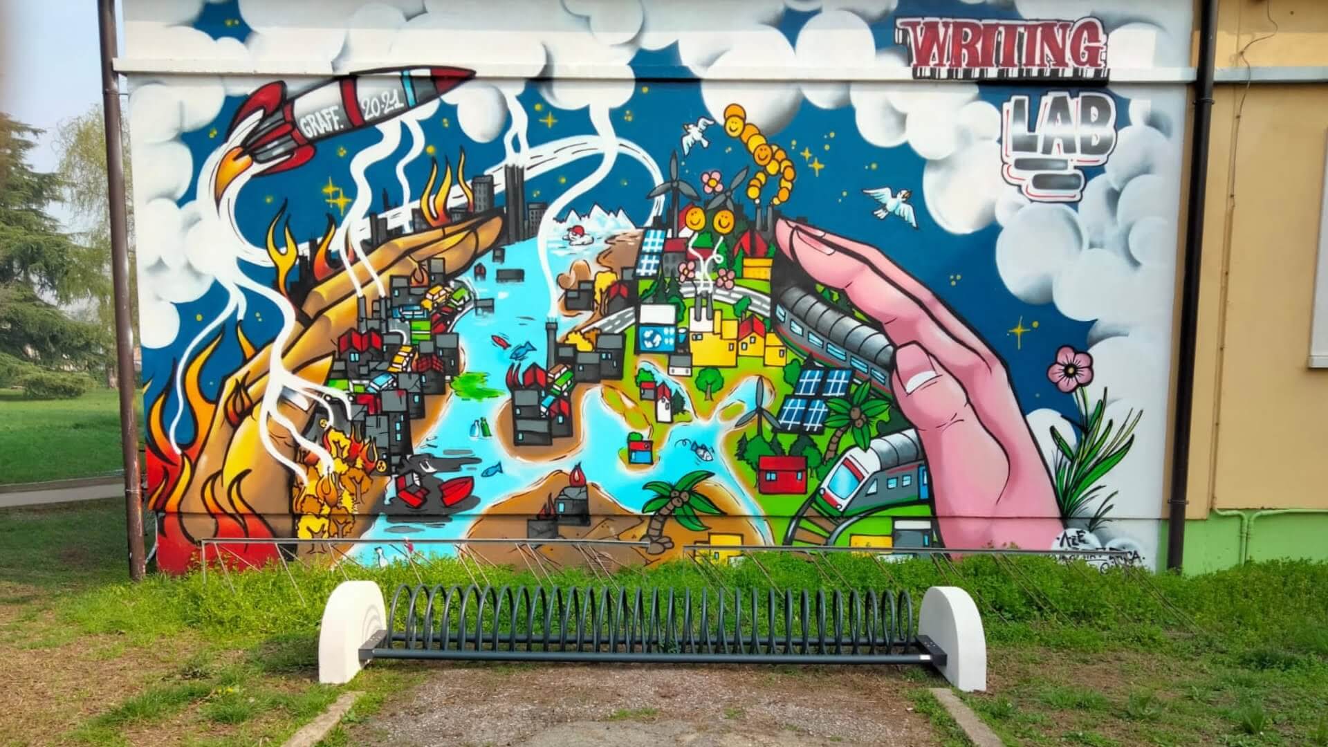 Spiraalvormig fietsenrek van staal met betonnen voeten, geplaatst voor een kleurrijke muurschildering in een groene omgeving.