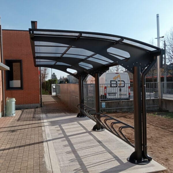 Fietsenstalling met zwart metalen overkapping en geïntegreerd fietsenrek op een betonnen ondergrond bij een bedrijfsgebouw.