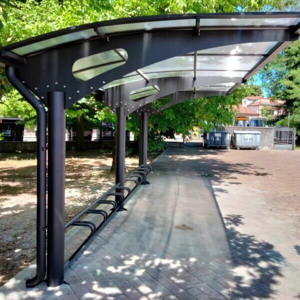 Robuuste rijwieloverkapping kopen voor stations Robuuste fietsenstalling met overkapping van staal en geïntegreerd fietsenrek, geplaatst op een betegeld plein met veel groen.