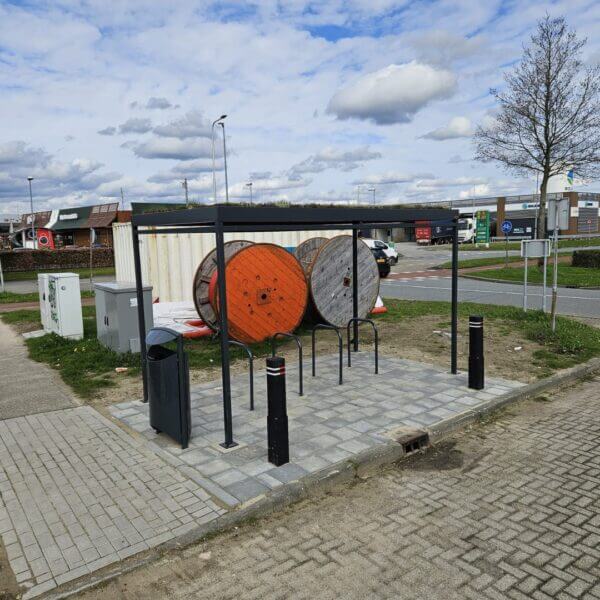 fietsenstalling-met-fietsnietjes-kopen Open fietsoverkapping met fietsnietjes en prullenbak naast twee houten kabelhaspels op een bedrijventerrein.