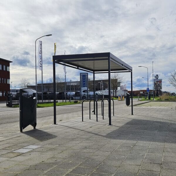 rijwielabri-direct-kopen-bij-umfeldt Vrijstaande rijwieloverkapping met vijf zwarte fietsnietjes op een ruim betegeld plein naast een bedrijventerrein. De overkapping heeft een strak zwart frame en een licht dak dat schaduw werpt op de parkeerplek.