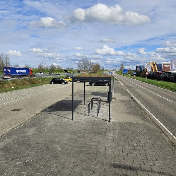 rijwieloverkapping-aanschaffen-reitdiep-fietsoverkapping-umfeldt Rijwieloverkapping met sedumdak en vijf zwarte fietsnietjes langs een industriële weg met parkeerplaatsen en voorbijrijdend verkeer op de achtergrond. De overkapping staat op een betegeld trottoir naast een afvalbak.