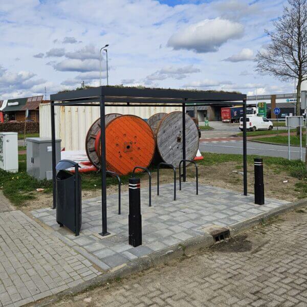 Rijwieloverkapping 4x2m direct online aanschaffen Rijwieloverkapping met fietsnietjes op een bestrate ondergrond, voorzien van een groen sedumdak. Op de achtergrond staan twee grote houten kabelhaspels en diverse voertuigen bij een bedrijventerrein.