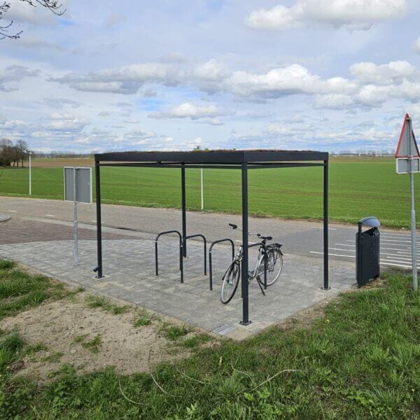 Rijwieloverkapping 4x2m met fietsnietjes direct bestellen Fietsoverkapping met sedumdak en vijf fietsnietjes aan de rand van een landelijke weg met uitzicht op een uitgestrekt groen veld. Eén fiets is geparkeerd onder de overkapping en er staat een afvalbak naast het geheel.