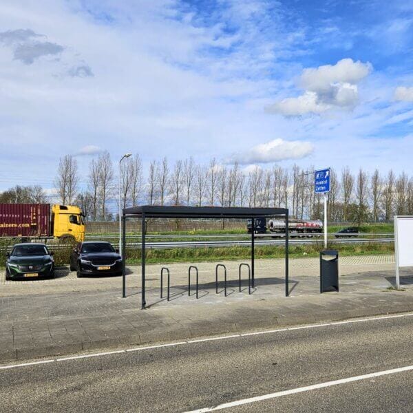 rijwieloverkapping-fietsnietjes-reitdiep-aanschaffen-bij-umfeldt Rijwieloverkapping met vijf zwarte fietsnietjes op een parkeerplaats langs de snelweg bij afrit Andelst Zetten, met passerende vrachtwagens en auto's op de achtergrond.