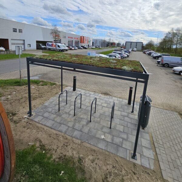 rijwieloverkapping-met-sedumdak-aanschaffen-online Fietsoverkapping met groen sedumdak en zwarte fietsnietjes als fietsenrek, geplaatst op een betegelde ondergrond naast een parkeerterrein op een bedrijventerrein.