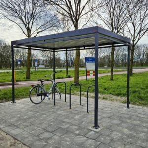 Rijwieloverkapping met metalen fietsnietjes als fietsenrek, geplaatst op een bestrate ondergrond naast een groenstrook met bomen. Onder de overkapping staat een enkele fiets geparkeerd.