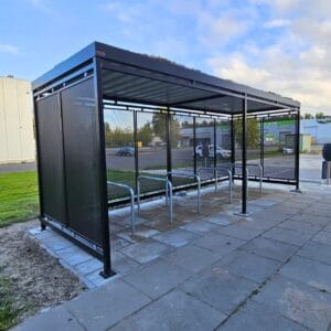 Rijwieloverkapping met vaste fietsenrekken en geïntegreerde oplaadpunten voor elektrische fietsen. Onder de metalen overkapping staan verschillende fietsen gestald, waaronder e-bikes en een scooter.