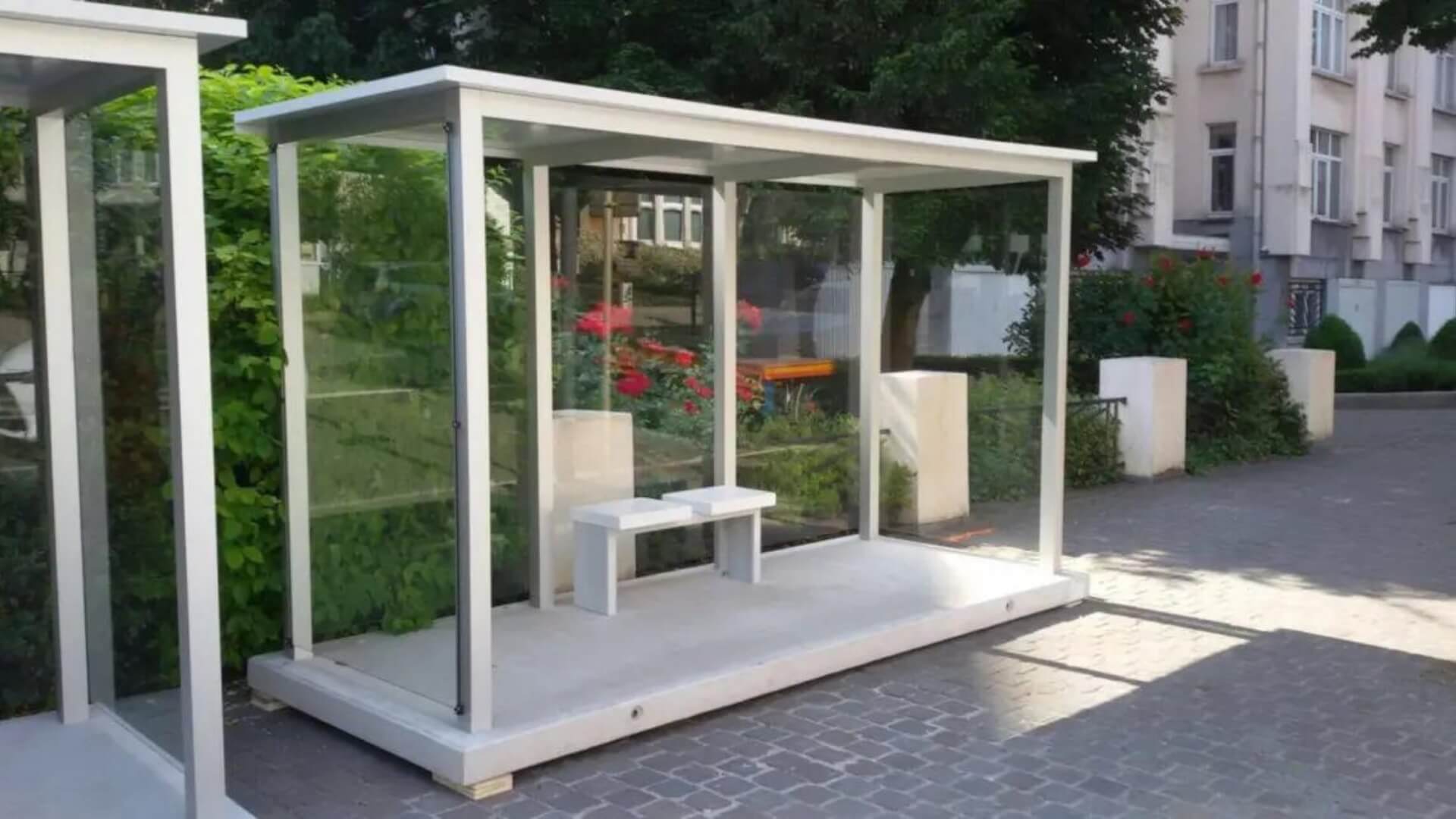 Moderne rookoverkapping met glazen wanden, zitbankje, vrijstaand geplaatst op een betonplaat op straat.