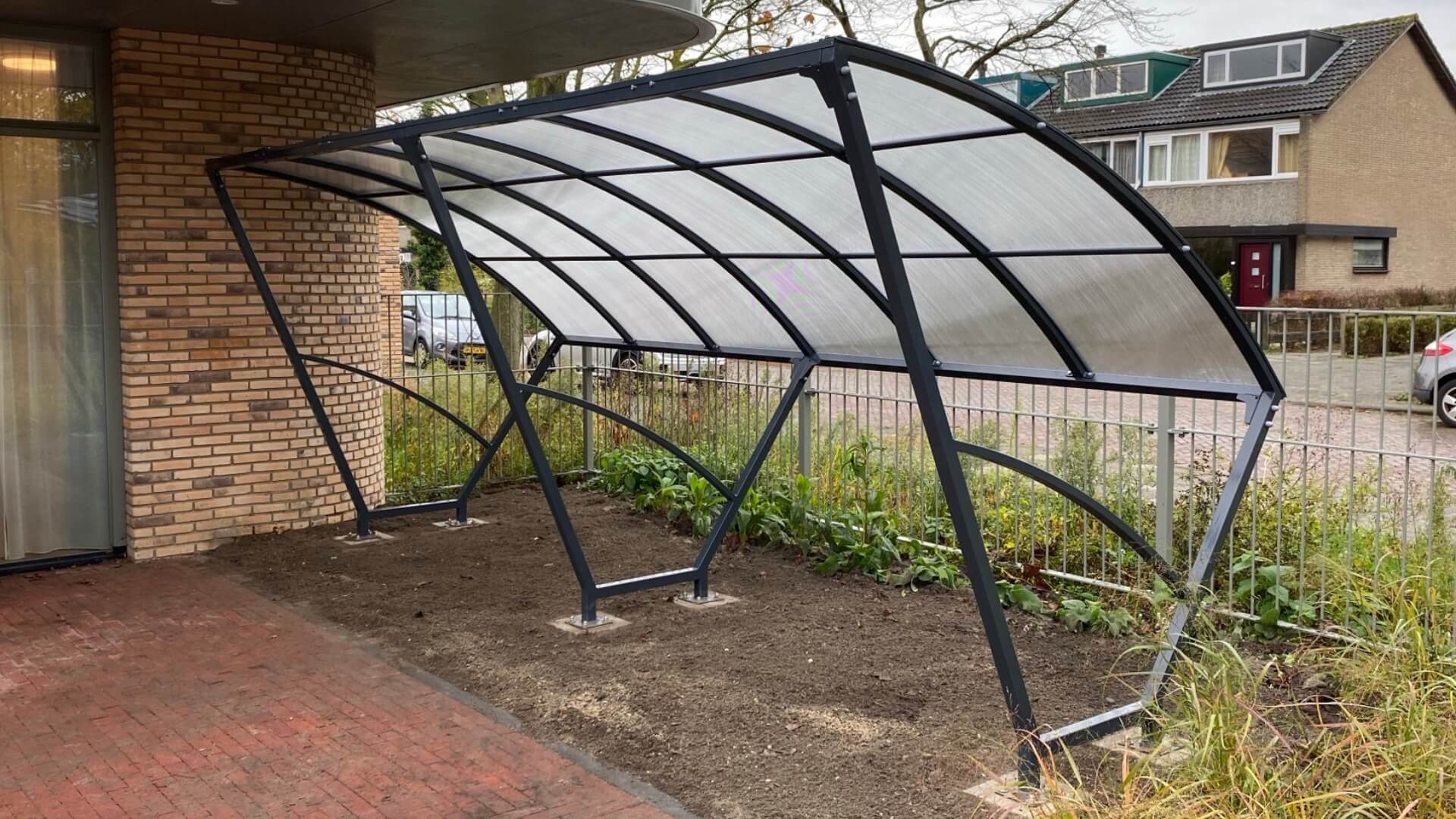 Moderne fietsoverkapping van 5 meter breed met gebogen transparant dak en stevig stalen frame, geschikt voor meerdere fietsen.