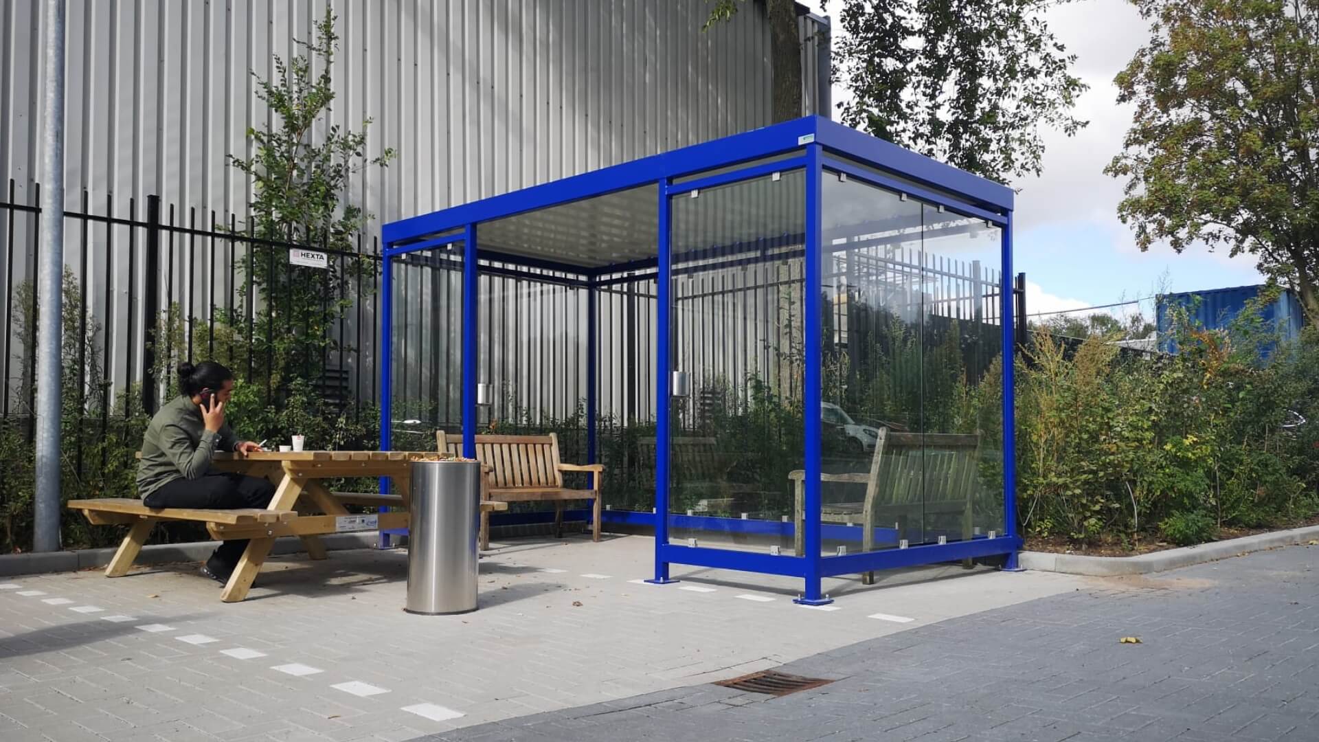Prefab rookoverkapping Keulen van Umfeldt met glazen wanden en blauw stalen frame, geplaatst bij bedrijfsterrein met buitenbanken en afvalbak