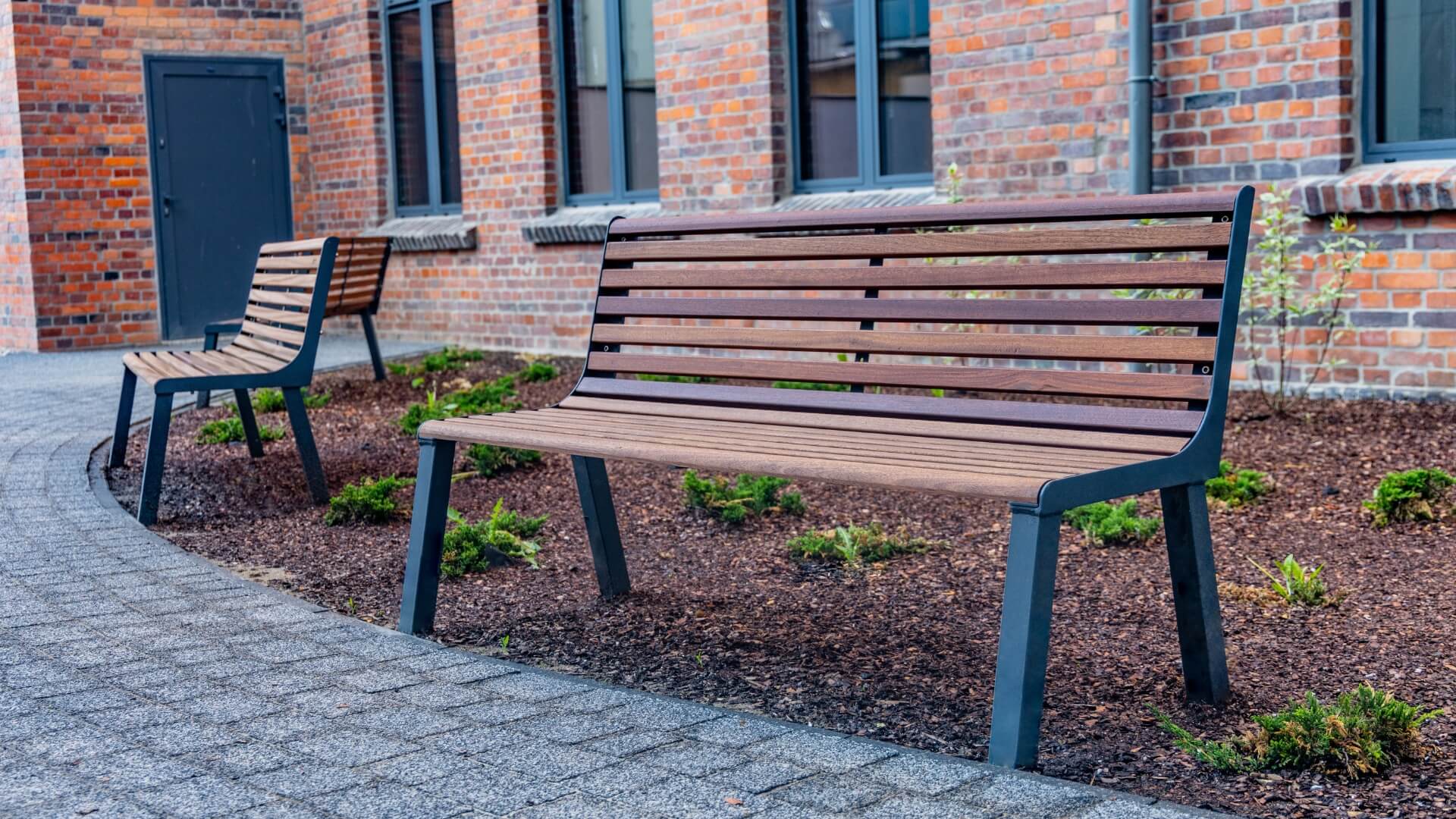 Houten parkbank met stalen frame in openbare buitenruimte bij gebouw, geschikt als duurzame buitenbank voor pleinen en parken.