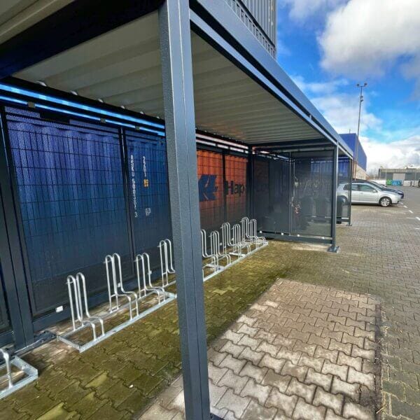 Functionele fietsoverkapping met rookruimte bestellen Donkergrijze fietsoverkapping naast een stapel zeecontainers, voorzien van een dubbele rij fietsenrekken met hoog-laag systeem en zijpanelen van geperforeerd staal.