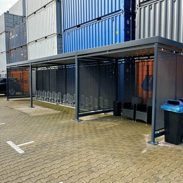 Fietsoverkapping en rookabri met strak design online kopen Zwarte fietsoverkapping met geïntegreerde fietsenrekken en een aparte zitruimte, geplaatst tegen een wand van gestapelde zeecontainers.