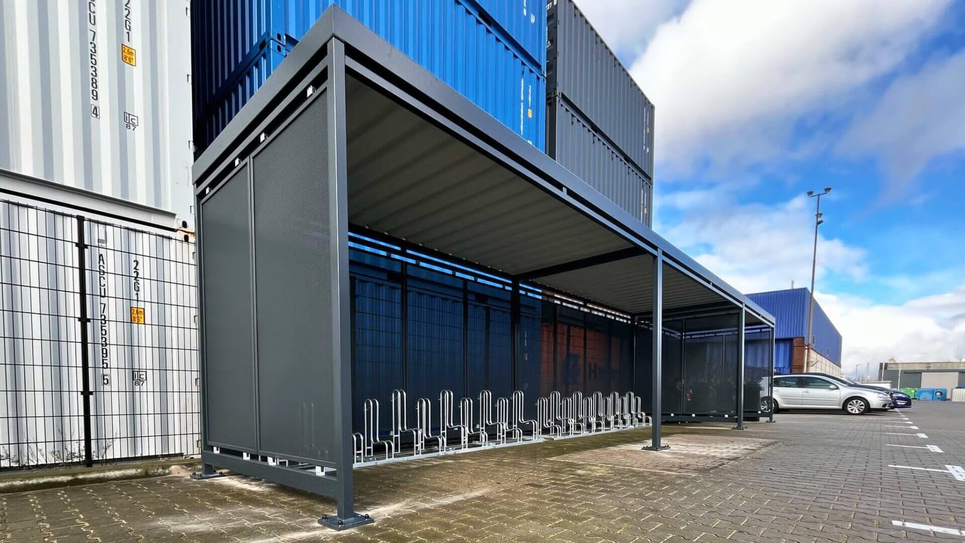 Grote stalen fietsoverkapping met geïntegreerde fietsenrekken, geplaatst op een industrieterrein naast stapels zeecontainers en parkeerplaatsen.
