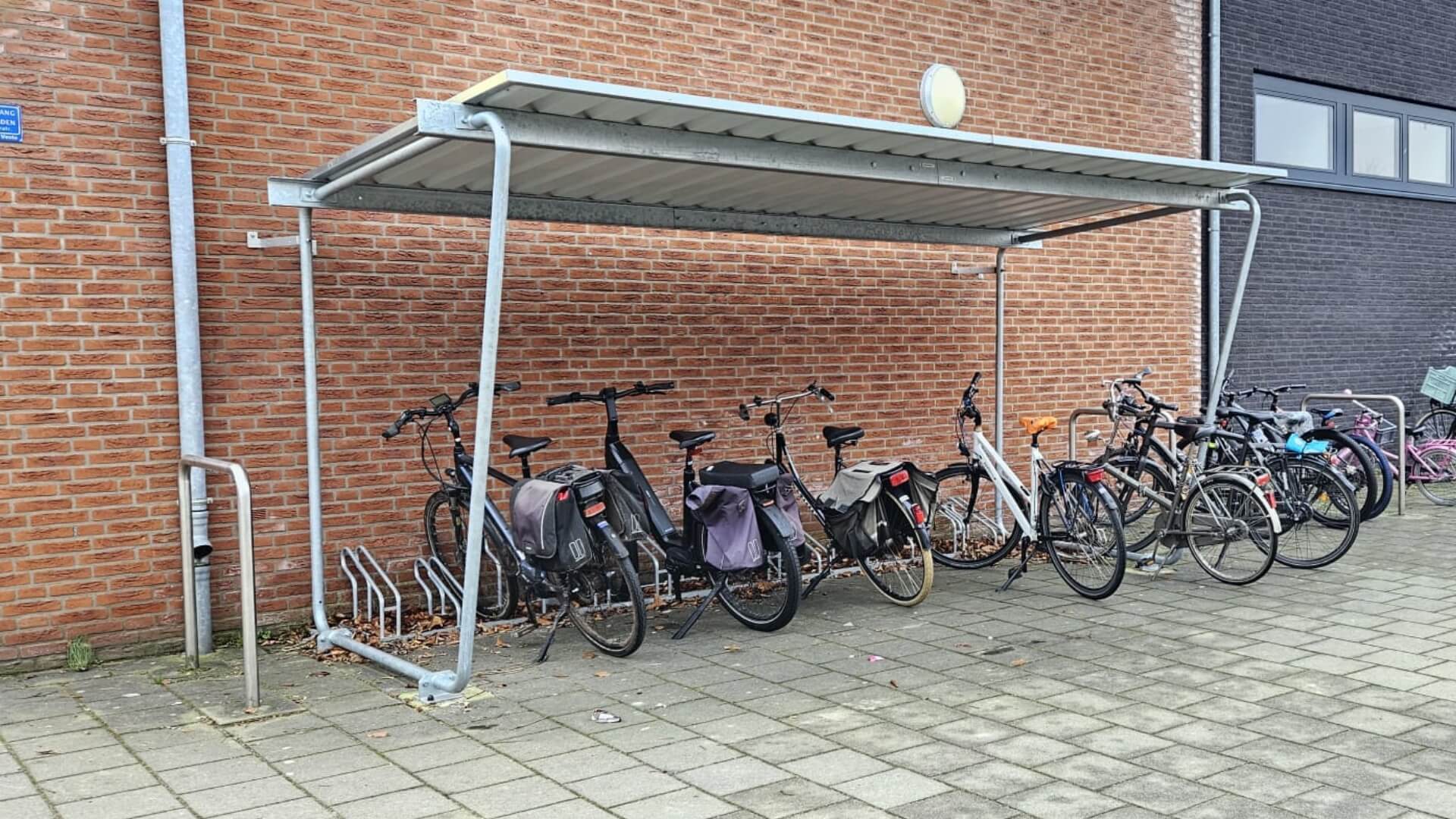 Bouwpakket fietsenstalling met metalen dak en fietsenrekken tegen een bakstenen muur van een school.