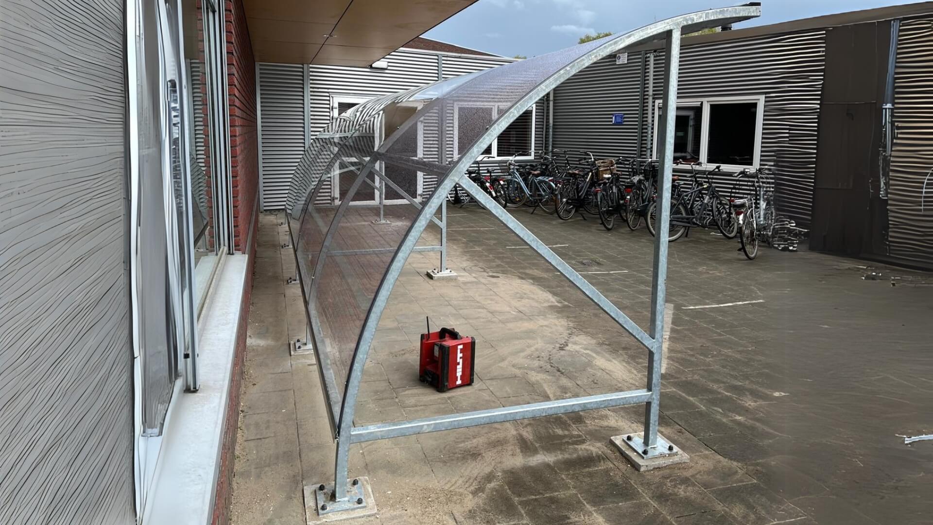 Grote bouwpakket fietsoverkapping met transparant dak en stalen frame op betonpoeren bij een school.