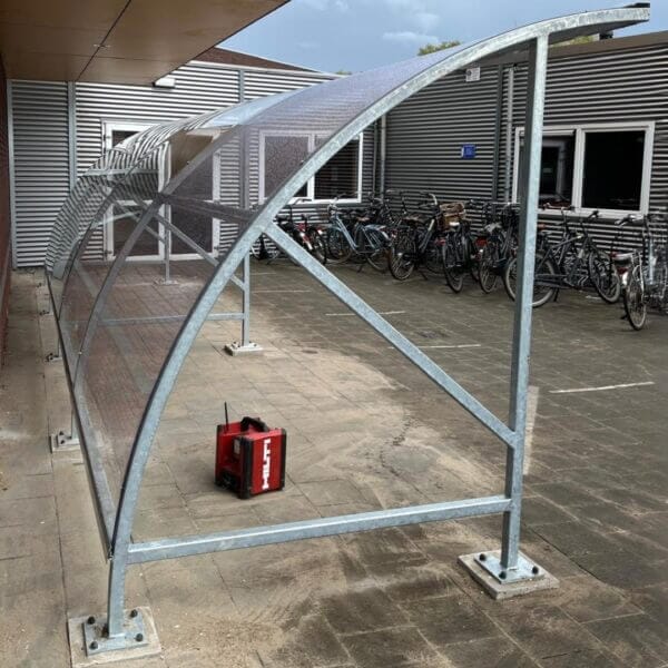 Bouwpakket fietsenstalling bestellen Bouwpakket fietsenstalling met transparant dak en gegalvaniseerd frame geplaatst op betonpoeren.