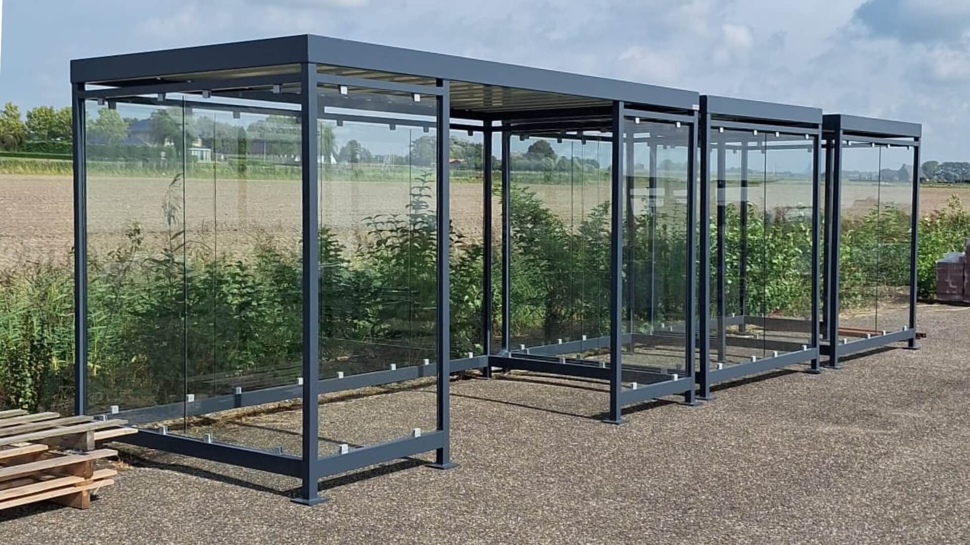 Drie gekoppelde overkappingen van staal en glas, geschikt als rookruimte of fietsoverkapping, geplaatst op een open terrein.