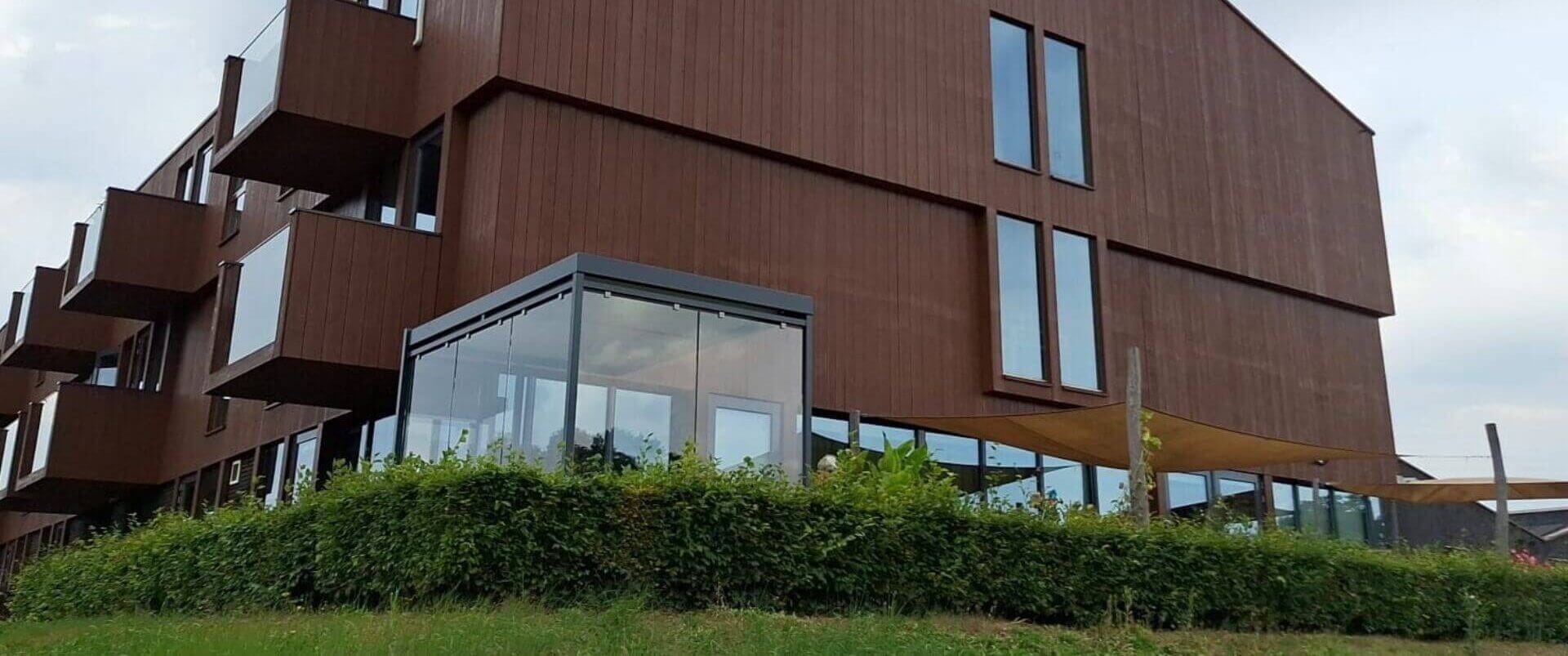 Glazen rookruimte naast modern houten zorggebouw, geplaatst op verhoogde locatie met omliggende haag bij een zorginstelling in Apeldoorn.