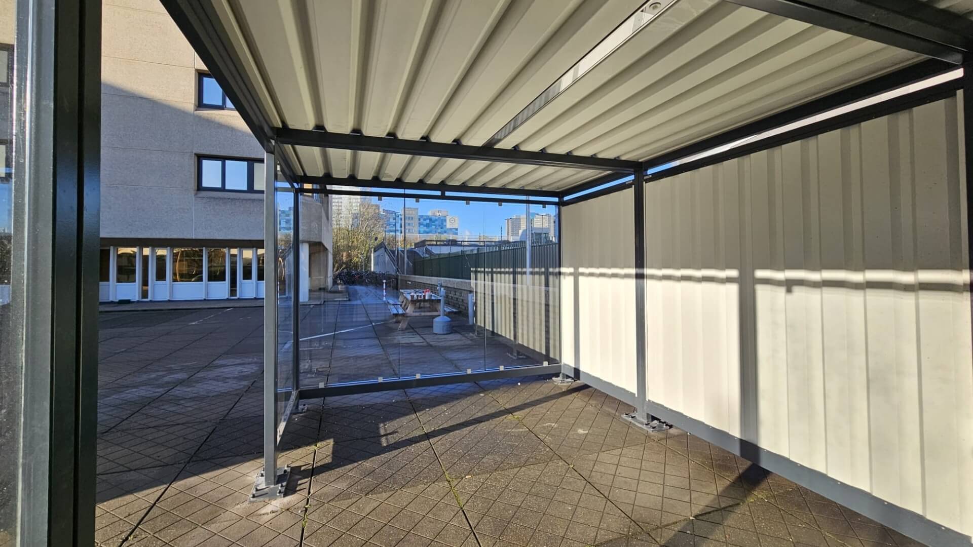 Maatwerk rookabri met glazen voorwand en trapeziumplaten wanden, geplaatst op het buitenterrein bij COA Amsterdam.